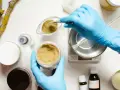Ręce w niebieskich rękawiczkach odmierzają proszek do wagi, przygotowując składniki kosmetyczne, w tym butylene glycol.