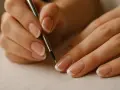 Dłonie z perfekcyjnym francuskim manicure, jedna z nich trzyma cienki pędzelek, jakby tworzyła dzieło sztuki.