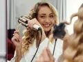 Uśmiechnięta blondynka w szlafroku pokazuje, jak zrobić fale prostownicą. Jej włosy są już częściowo pofalowane.