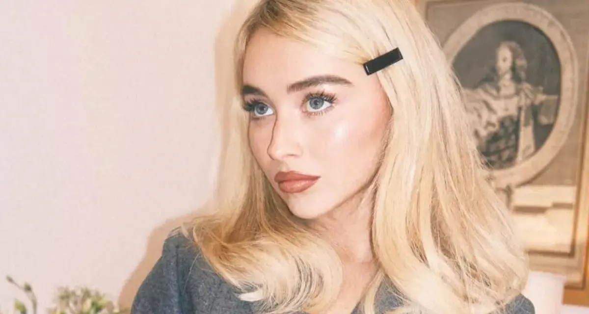 Blondynka z **side part fryzurą** i czarną spinką, w szarym żakiecie, z błękitnymi oczami.
