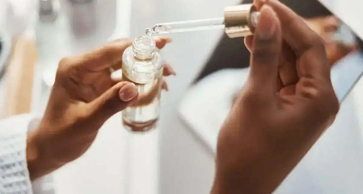 Dłonie trzymają buteleczkę z serum. Kropla z pipety, zawierająca kwas ferulowy, spada do buteleczki, przygotowując pielęgnację.