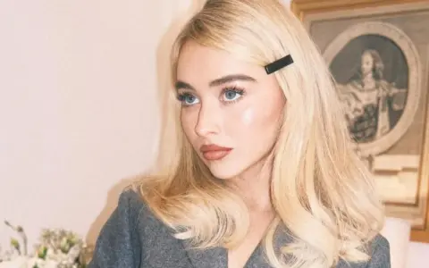 Blondynka z **side part fryzurą** i czarną spinką, w szarym żakiecie, z błękitnymi oczami.