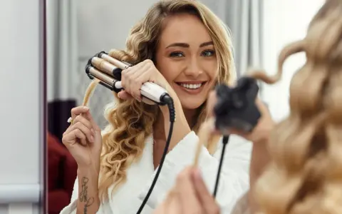 Uśmiechnięta blondynka w szlafroku pokazuje, jak zrobić fale prostownicą. Jej włosy są już częściowo pofalowane.