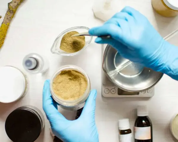 Ręce w niebieskich rękawiczkach odmierzają proszek do wagi, przygotowując składniki kosmetyczne, w tym butylene glycol.