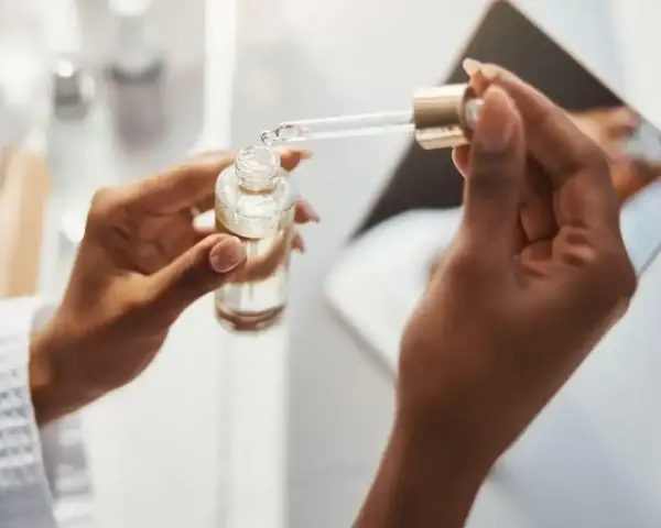 Dłonie trzymają buteleczkę z serum. Kropla z pipety, zawierająca kwas ferulowy, spada do buteleczki, przygotowując pielęgnację.