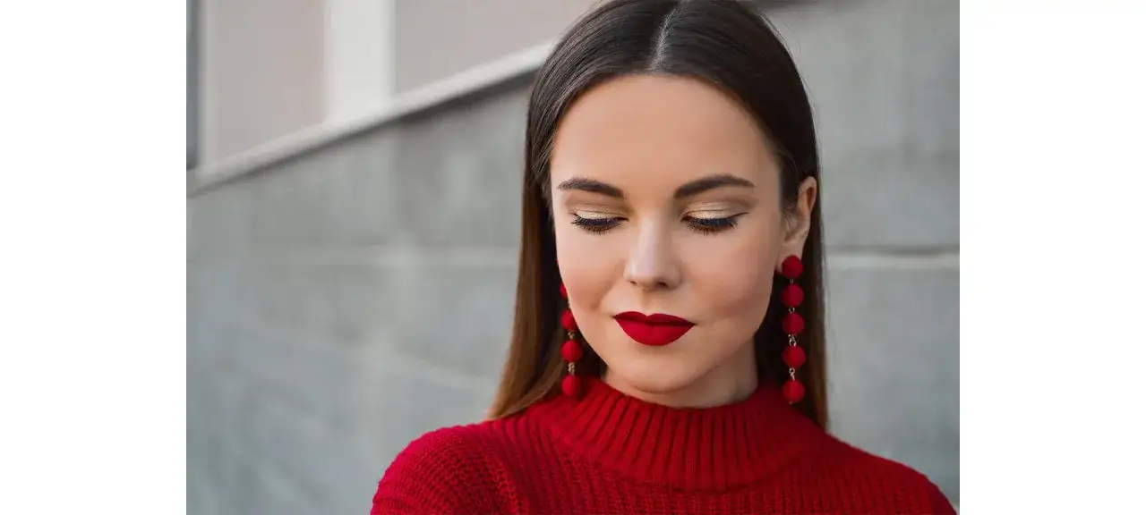 Elegancka brunetka w czerwonym swetrze i długich, czerwonych kolczykach, z idealnym makijażem.