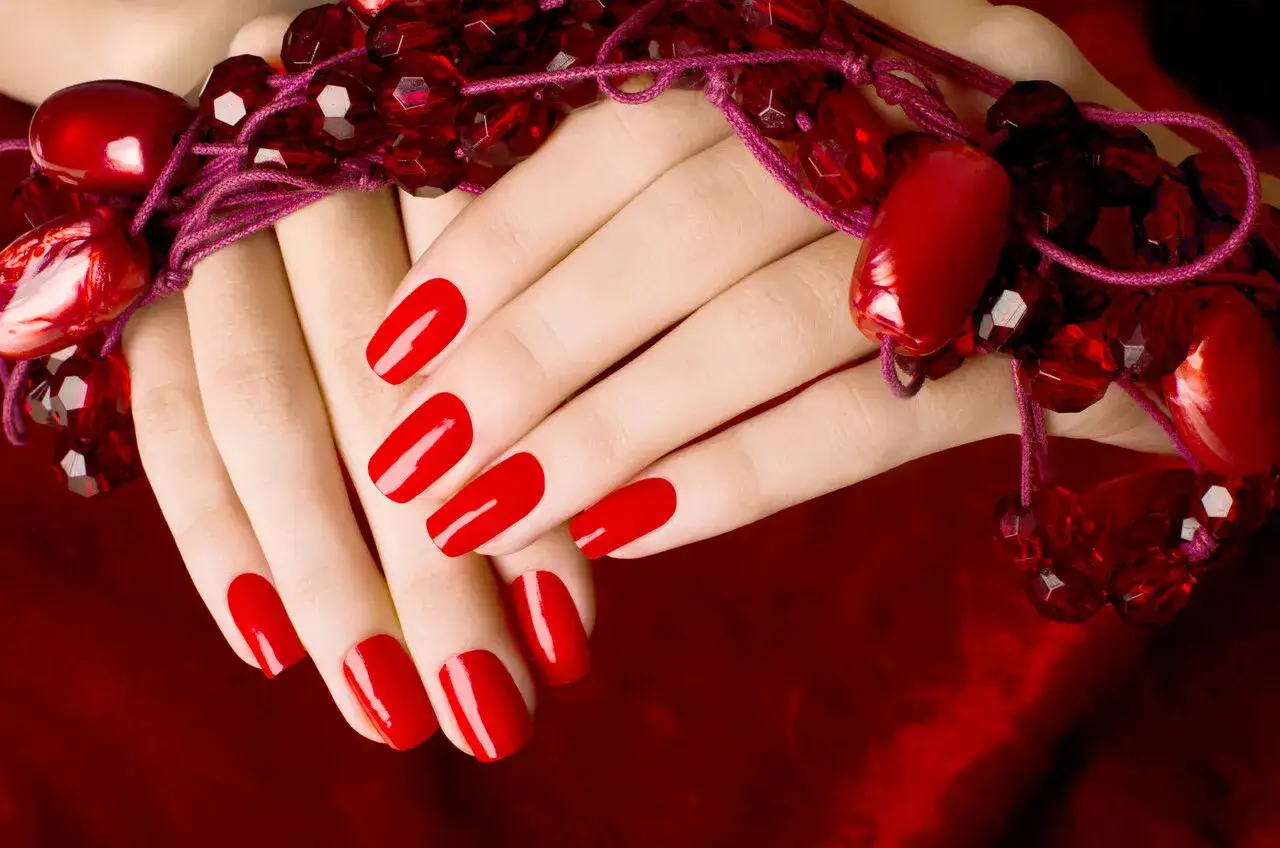 Dłonie z idealnym, błyszczącym manicurem w kolorze najmodniejszych czerwonych paznokci, ozdobione koralikami.