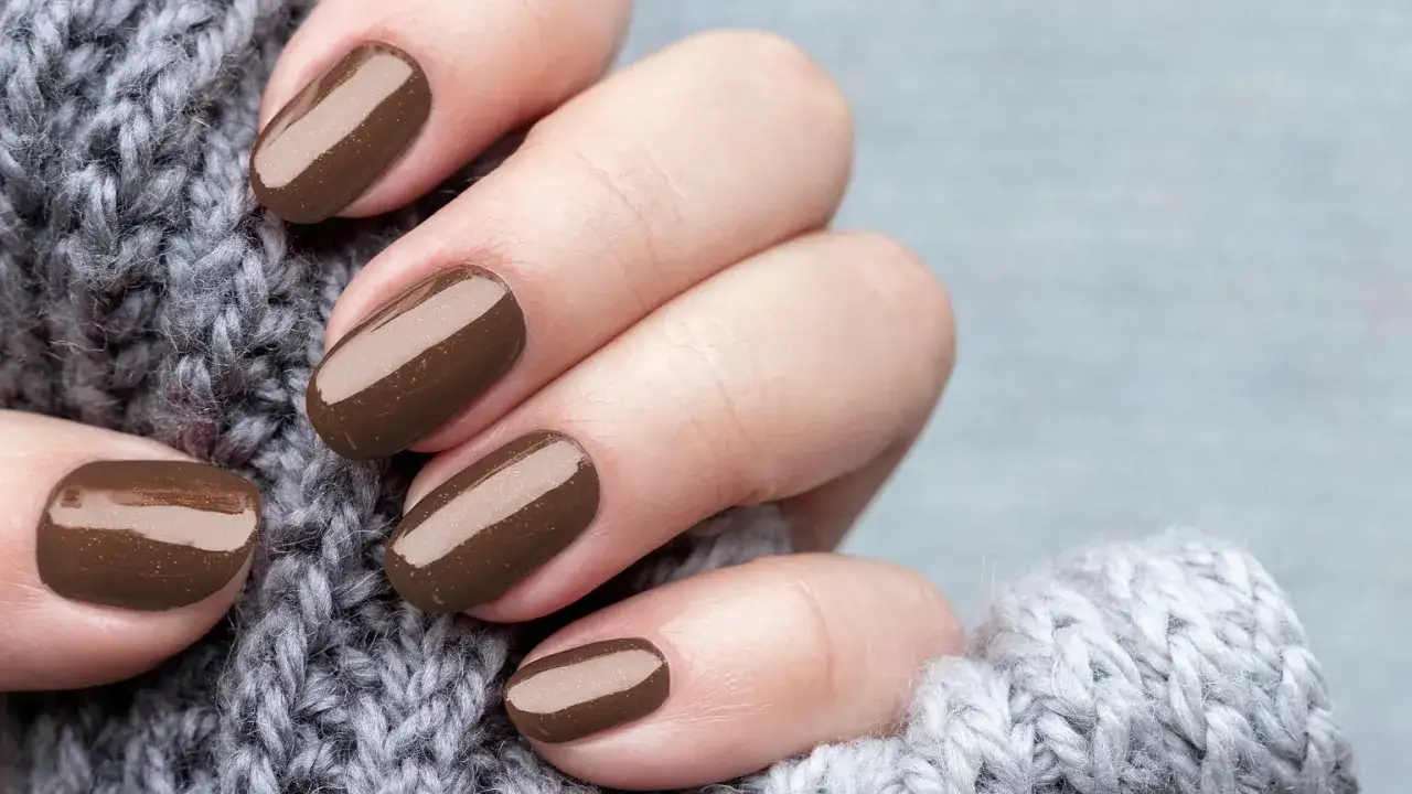 Dłonie z perfekcyjnym manicurem kombinowanym w odcieniu gorzkiej czekolady, otulone miękkim, szarym swetrem.
